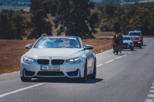 White convertible car driving on a road with cyclists.